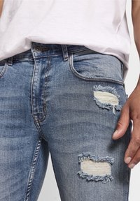 Jeans de mezclilla azul claro con parches desgastados en los muslos, con bordes deshilachados. Diseño clásico de cinco bolsillos con cierre de botón.