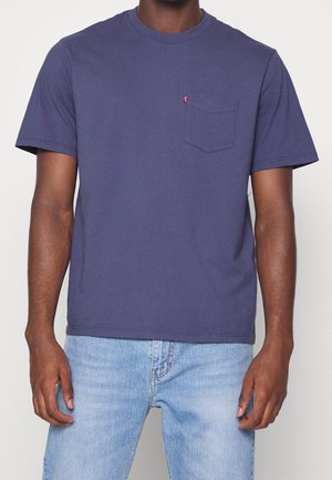 Homme portant un t-shirt uni bleu foncé à manches courtes avec une poche poitrine et un jean bleu clair, debout devant un fond uni.