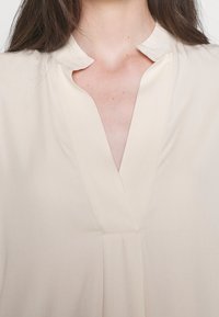 Femme portant un chemisier beige clair à col en V avec un petit col montant, montrant son cou et le haut de sa poitrine.