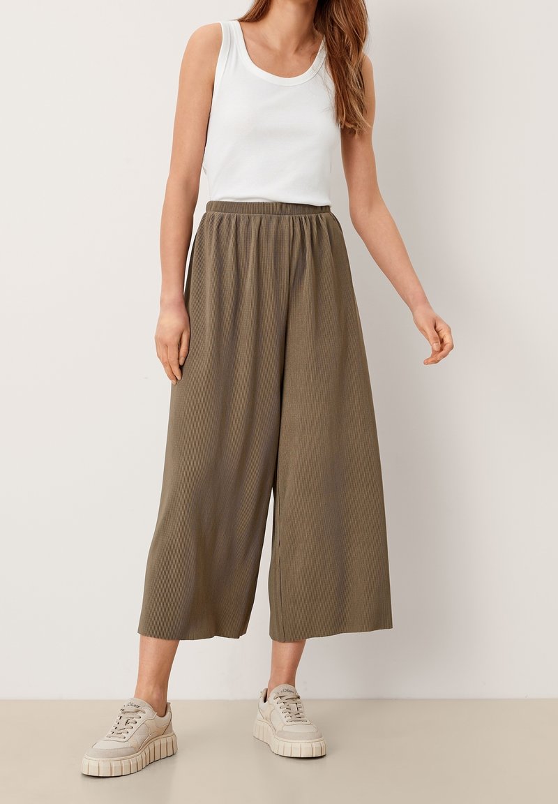 Femme portant un haut blanc sans manches, un pantalon large olive coupé court et des baskets compensées beige, debout devant un fond clair uni.