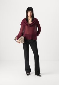 Top trasparente bordeaux con scollatura drappeggiata, maniche lunghe e pantaloni neri a palazzo. Tiene una borsa metallica argento con design plissettato.
