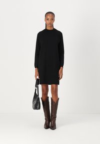 Melnas garās piedurknes kleita, līdz ceļiem, ar ribotu apkakli, saskaņota ar brūniem kovboju zābakiem un mazu melnu rokassomu.