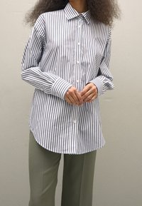 Persona che indossa una camicia bianca a maniche lunghe con strisce verticali navy e pantaloni verde chiaro, in piedi contro un muro semplice di colore chiaro.