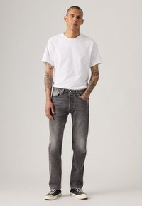 Man met tatoeages in een simpele witte t-shirt, vervaagde zwarte jeans en zwarte lage sneakers, staand tegen een effen achtergrond.