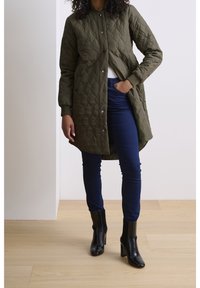 Kurtka pikowana w oliwkowym kolorze z zapięciem na guziki i prążkowanymi mankietami, w zestawieniu z ciemnoniebieskimi rurkami i eleganckimi czarnymi botkami na obcasie.