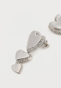 Ασημένιο κλιπ σε σχήμα καρδιάς με τρία μεγέθη, διαθέτει λεία επιφάνεια, διακοσμημένο με στρας στα άκρα. Εγχάρακτο το όνομα της μάρκας "GUESS".