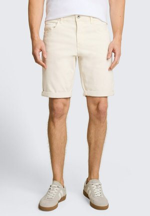 Mann trägt beige Shorts mit umgeschlagenem Saum und hellgraue Turnschuhe, steht vor weißem Hintergrund.
