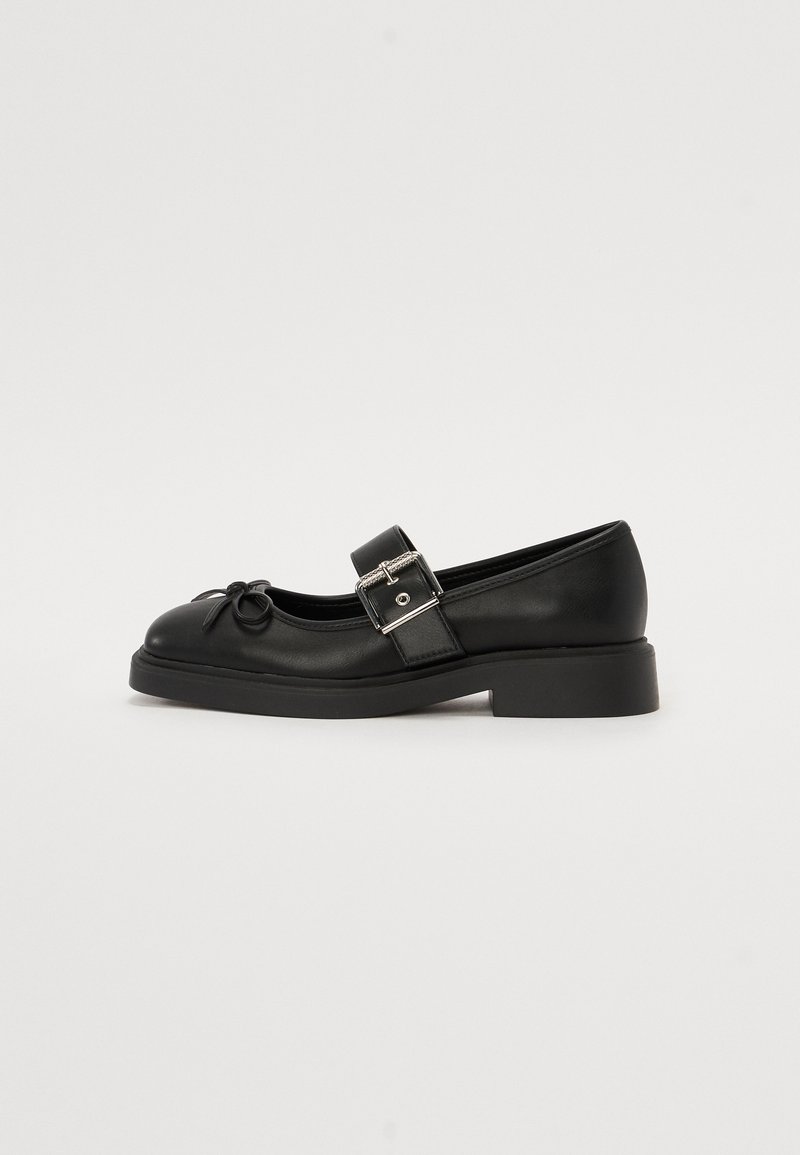 Black leather Mary Jane shoe with a round toe, ankle strap, and silver buckle detailing. Features a flat, chunky sole and bow accent.