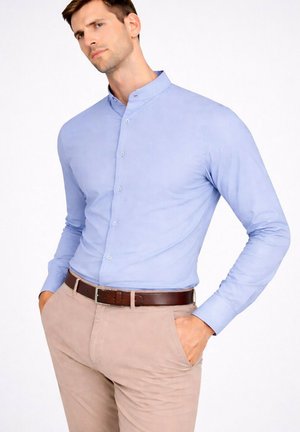 Homme portant une chemise bleu clair à col mandarin rentrée dans un pantalon beige avec une ceinture en cuir marron, mains dans les poches, fond neutre.