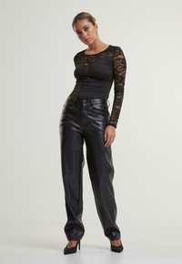 Zwarte bodysuit met lange mouwen en kanten accenten; hoog-wijze nepleder broek; model draagt zwarte hoge hakken en poseert zelfverzekerd.
