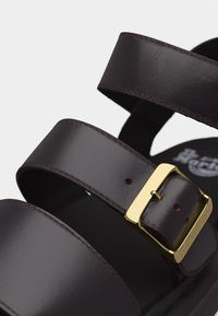 Σκούρο καφέ δερμάτινο σανδάλι με φαρδιές λουρίδες και χρυσή αγκράφα χαραγμένη με το όνομα της μάρκας, δείχνοντας λεπτομερείς ραφές και ματ φινίρισμα.