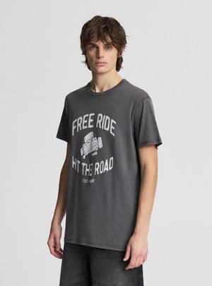 Młody mężczyzna z ciemnymi falowanymi włosami, ubrany w wyblakłą czarną koszulkę z napisem "FREE RIDE HIT THE ROAD" i grafiką deskorolki.