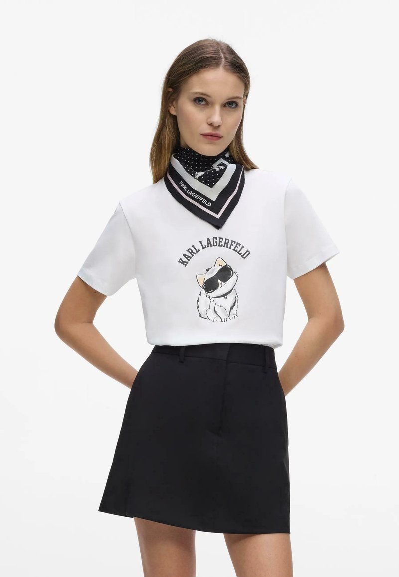Frau trägt ein weißes T-Shirt mit einem Katzenmotiv und dem Schriftzug "KARL LAGERFELD", einen schwarzen Rock und einen schwarzen gepunkteten Schal um den Hals.