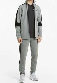 Chaqueta deportiva gris con acentos negros, cierre delantero y pantalones gris ajustados. Combinada con zapatillas gris, que presentan un diseño sencillo con un logo.