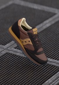 Brun og svart blandet materiale sneakers med gule detaljer, tykke snøringer og en teksturert såle. Har synlig merking på tungen.