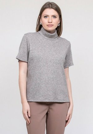 Jeune femme aux cheveux bruns lisses portant des boucles d'oreilles argentées en forme d'anneau, un pull à col roulé gris à manches courtes et un pantalon marron clair, debout.