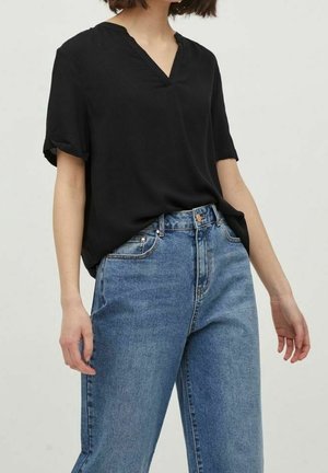 Kvinde iført en sort kortærmet V-hals bluse, stoppet ned i blå højtaljede denimjeans, stående mod en ensfarvet baggrund.