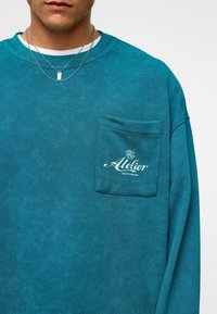 Teal, langarm sweatshirt gemaakt van zacht materiaal met een voork pocket waarop de witte tekst "Atelier" is gedrukt. Gestructureerd oppervlak en een ontspannen pasvorm.