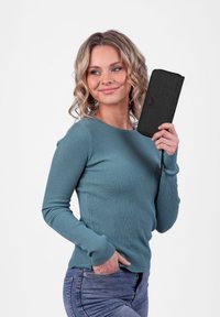 Black fabric wallet with a textured finish, featuring a zipper, held by a person in a teal ribbed long-sleeve shirt and blue jeans.