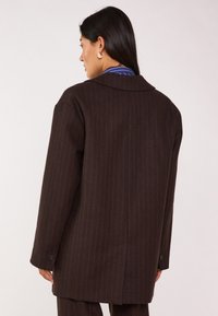 Blazer a righe marrone scuro con colletto a punta, maniche lunghe e dettagli con bottoni. Il tessuto ha una finitura fine e una forma strutturata.