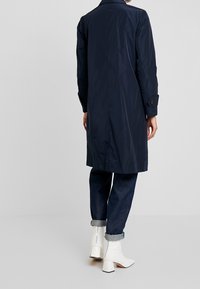 Granatowy wodoodporny płaszcz trench z prostym fasonem, zapięciem na guziki przy rękawach oraz średniej długości. Połączony z ciemnymi dżinsami i białymi botkami na obcasie.