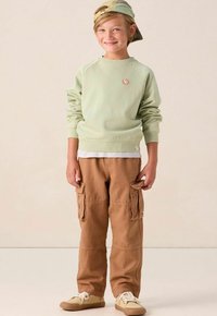 Glunderend kind dat een groene sweatshirt, bruine cargobroek, beige sneakers en een achterstevoren gedragen groene pet draagt, staande op een lichte achtergrond.