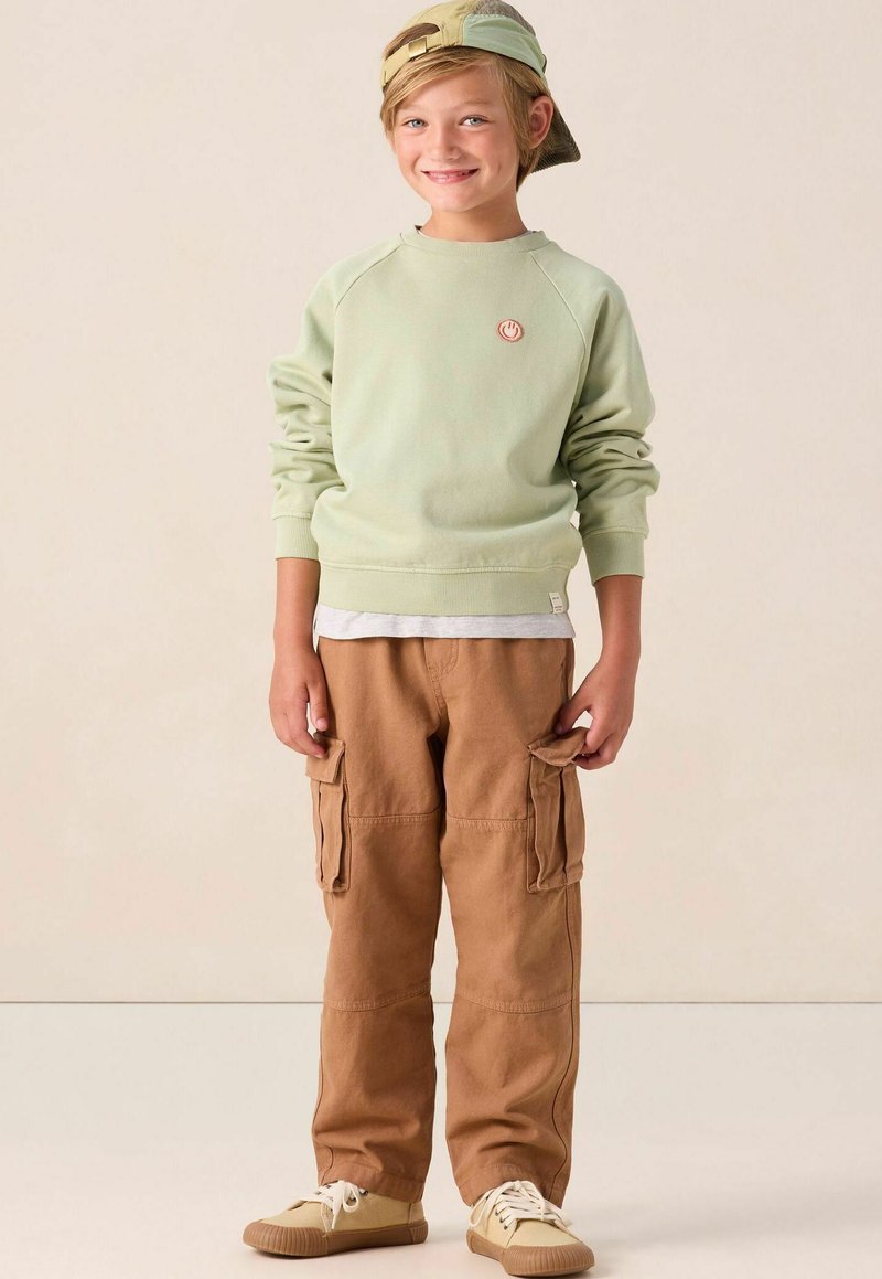 Glunderend kind dat een groene sweatshirt, bruine cargobroek, beige sneakers en een achterstevoren gedragen groene pet draagt, staande op een lichte achtergrond.