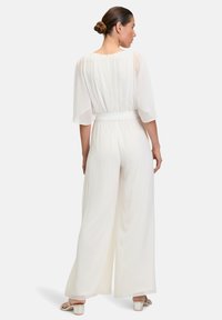 Fehér overál, áttetsző, fodros ujjaival és testhezálló derék résszel, széles szárú nadrágokkal és sima textúrával. Egyszerű és elegáns dizájn.