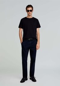 Sort bomulds t-shirt med rund hals, marineblå bukser, sort bælte og sorte sko. Modellen har solbriller på og poserer med hænderne i lommerne.
