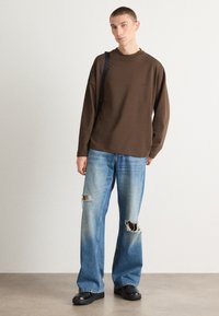 Chemise marron à manches longues, jeans en denim bleu taille ample avec des déchirures usées, et chaussures à plateforme noires, se tenant contre un fond neutre.