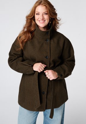 Femme souriante aux longs cheveux roux portant un manteau à carreaux marron foncé, boutonné et noué à la taille, avec un jean bleu clair.