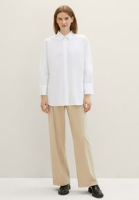 Chemise blanche à boutons avec un col pointu et des manches longues, associée à un pantalon beige à jambes larges et des chaussures noires, le tout sur un fond neutre.
