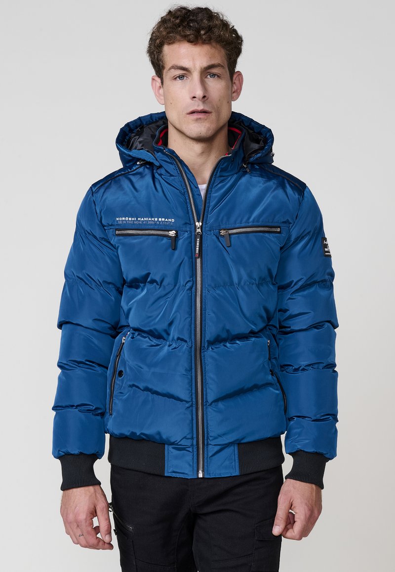 Giacca imbottita blu con chiusura a zip, tasche laterali e sul petto, polsini a coste e cappuccio. Il tessuto ha una texture liscia e lucida.
