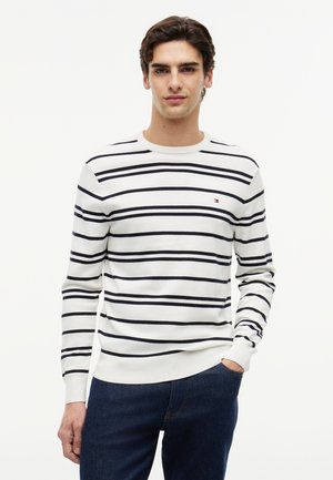 Ung mand iført en hvid langærmet skjorte med sorte vandrette striber og mørkeblå jeans, stående med den ene hånd i lommen.