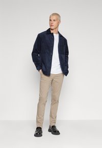 Navy corduroy shirt jacket with a button closure, paired with a white t-shirt, beige chinos, and black shoes. Simple, structured design.