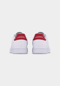 Hvite Lacoste-sneakers sett bakfra med røde hælfliker som viser Lacoste-merkevarenavn i hvit tekst.