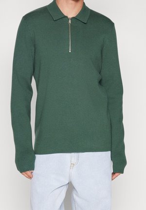Homme portant un polo à manches longues vert foncé avec une fermeture éclair à l'avant, associé à un jean bleu clair, debout devant un fond uni.