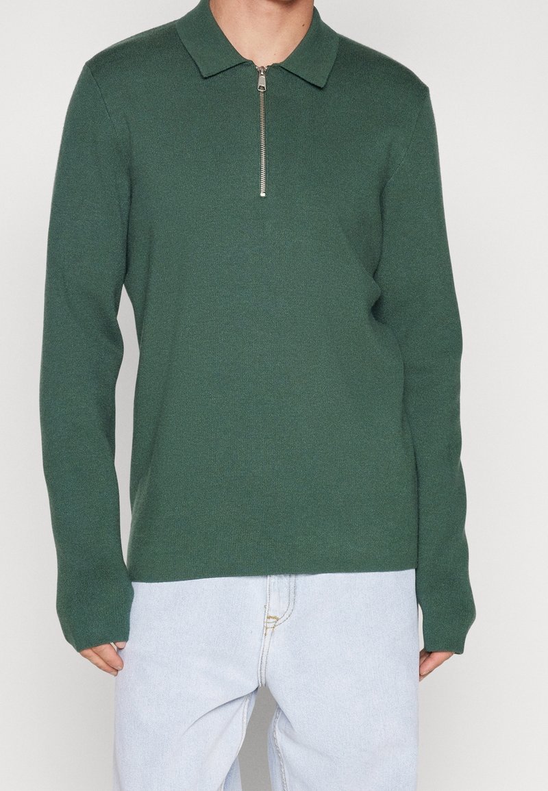 Homme portant un polo à manches longues vert foncé avec une fermeture éclair à l'avant, associé à un jean bleu clair, debout devant un fond uni.