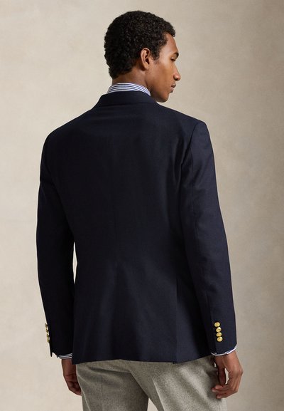 Blazer azul-marinho com corte ajustado, única fenda, botões dourados nos punhos e textura suave. Usado sobre uma camisa às riscas.