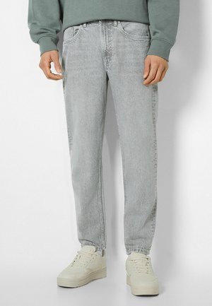 Mand iført lysegrå jeans, beige sneakers og en grøn sweatshirt står mod en hvid baggrund.