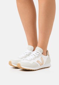 Hvite joggesko med teksturert mesh og semsket detaljer, med beige aksenter og gummisåle. Snøring med runde hvite lisser.
