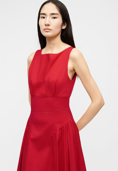 Γυναίκα με μακριά σκούρα μαλλιά, φορώντας αμάνικο κόκκινο φόρεμα με εφαρμοστή μέση και πλισέ φούστα, στέκεται μπροστά σε απλό ανοιχτό φόντο.