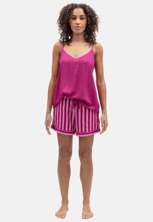 Donna con capelli ricci che indossa un top senza maniche rosa e pantaloncini rosa a righe, in piedi a piedi nudi su uno sfondo neutro.