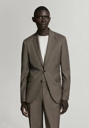 Hombre negro vestido con una chaqueta de traje marrón y pantalones a juego sobre una camisa blanca, de pie contra un fondo claro y liso.
