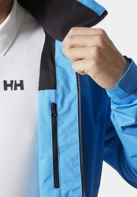 Chaqueta impermeable azul con detalles en negro, bolsillo con cremallera y cuello forrado de forro polar; llevada sobre una camisa blanca con textura que muestra un logo.