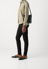 Persona con blusa beige de puños con volantes, pantalones negros ajustados, mocasines negros sin cordones, anillos de oro y bolso negro de hombro contra un fondo blanco.