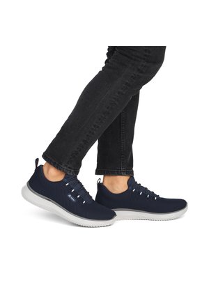 Person iført sorte jeans og marineblå mesh-sneakers med hvide såler, stående på en hvid baggrund.