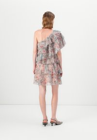 Kvinna som bär en ärmlös, enaxlad, lager-på-lager blommig klänning i pastellfärger, står med ryggen mot kameran, iförd lågklackade slingbacks.