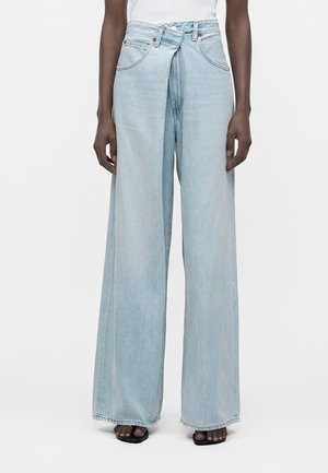 Osoba nosząca jasnoniebieskie szerokie dżinsy z wysokim stanem i nakładającymi się przednimi panelami, w połączeniu z czarnymi sandałami z odkrytymi palcami.