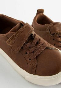 Brązowe syntetyczne sneakersy o fakturowanym wykończeniu, z sznurówkami i paskiem na rzep, uzupełnione białą gumową podeszwą.